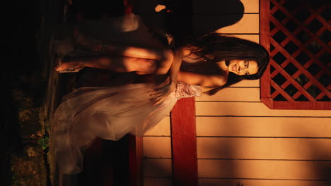 Slow-motion-vertical-handheld-shot-of-a-young-pretty-woman-dressed-in-a-pink-dress-sitting-in-front-of-a-wooden-house-with-red-colored-wood-while-she-looks-to-the-side-and-smiles-during-sunset