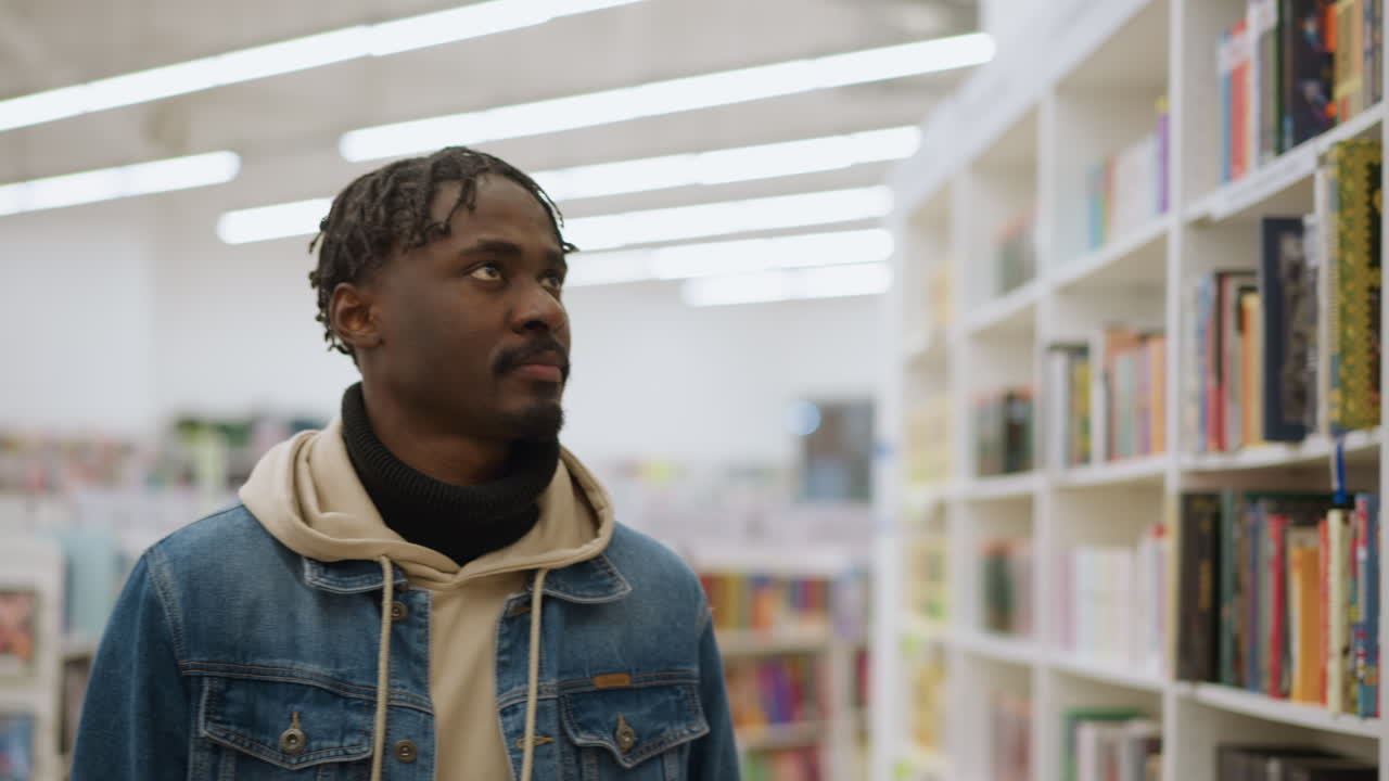 Young guy walking through bookstore, scanning bookshelves filled with colorful book spines. Soft indoor lighting highlights shelves and creates calm atmosphere. Focus on browsing books