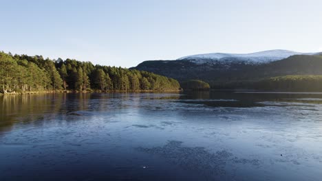 Drohnenaufnahmen-Aus-Der-Luft,-Die-Sich-über-Die-Oberfläche-Eines-Gefrorenen,-Eisbedeckten-Sees-Im-Cairngorms-Nationalpark-In-Schottland-Drehen,-Mit-Einem-Einheimischen-Wald-Aus-Schottischen-Kiefern-Entlang-Der-Küste-Und-Einem-Klaren-Blauen-Himmel