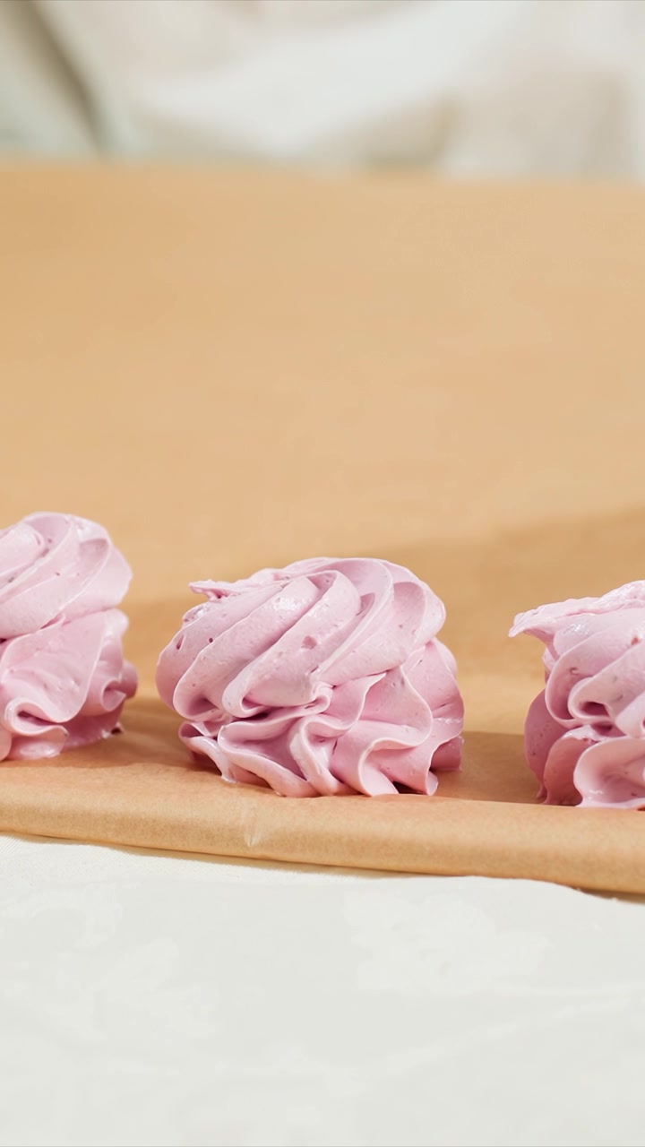 Close-up tiga kue meringue merah muda yang tersusun. Kue mempertahankan tekstur pusaran lembut dan warna pastel yang konsisten. Pengaturan dalam ruangan dengan latar belakang netral yang diterangi dengan lembut