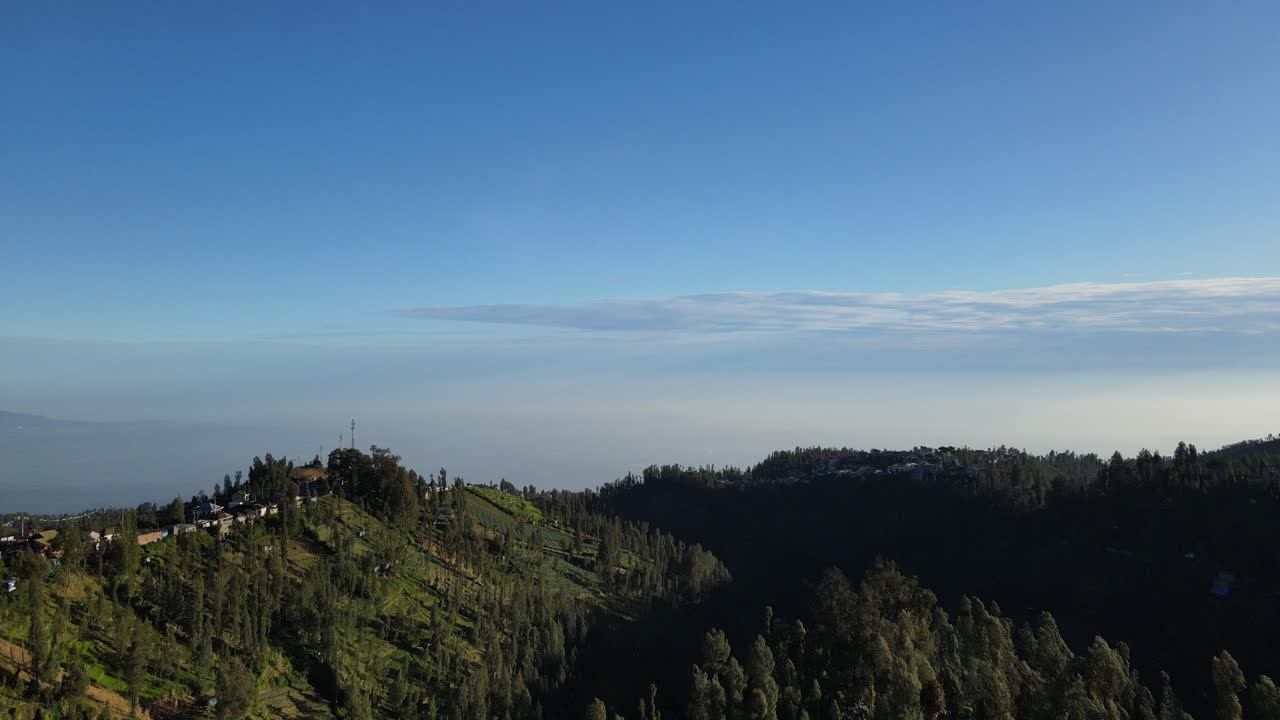 Aerial drone footage of Indonesian highland ridge with scattered houses, green slopes, pine trees, and wide horizon under bright clear blue sky