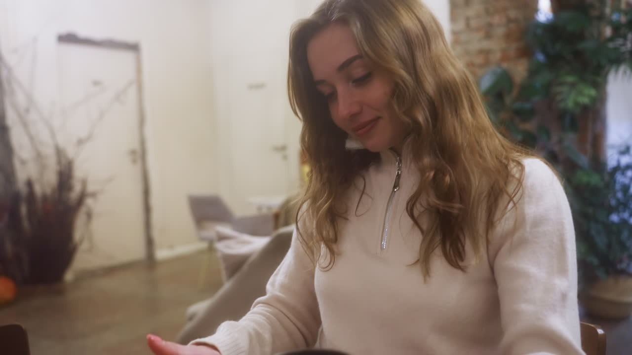 Cheerful girl in beige shirt holding coffee cup, smiling while sitting on chair in cozy cafe. Relaxed vibe with soft background and natural lighting enhancing inviting atmosphere