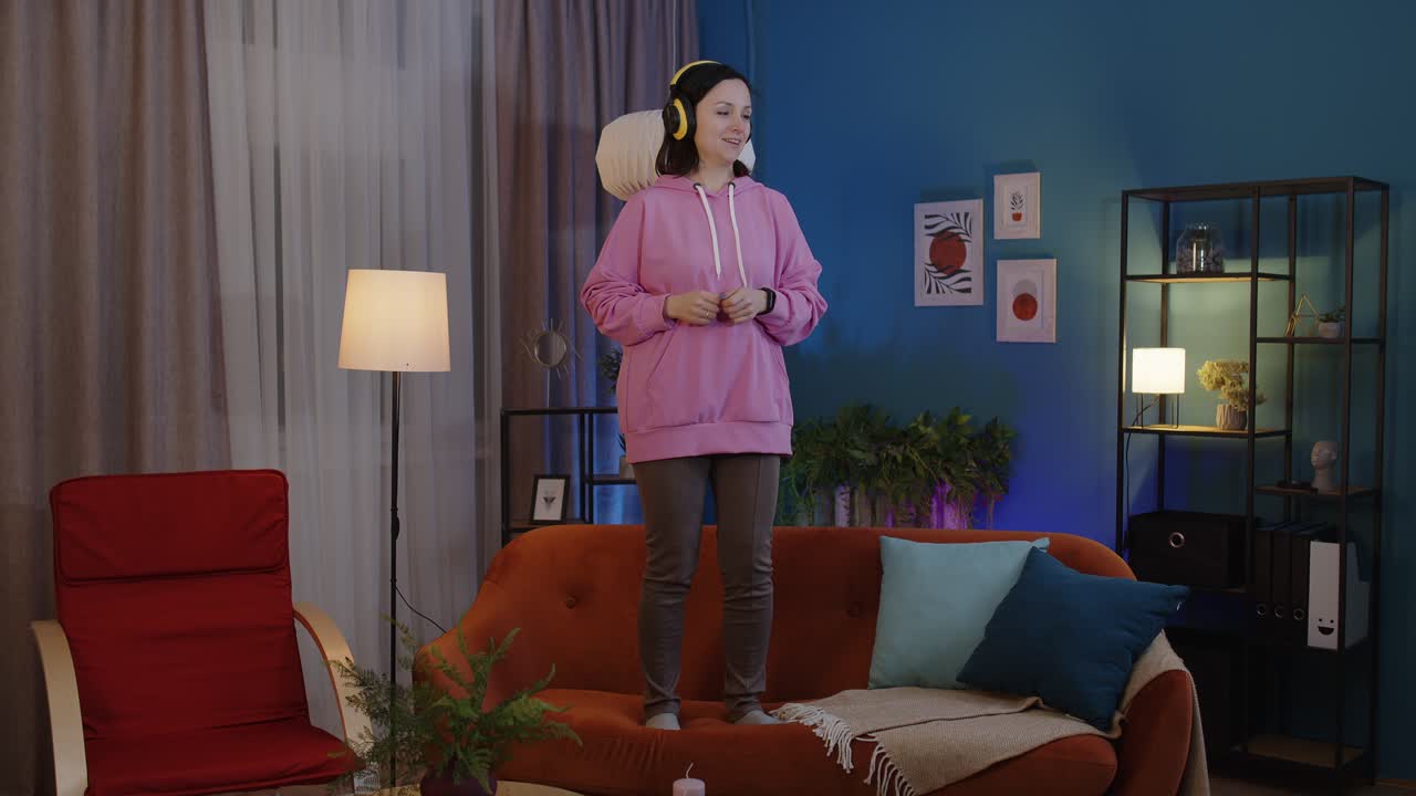mujer adulta feliz con auriculares bailando y saltando mientras escucha música en casa sola en el sofá