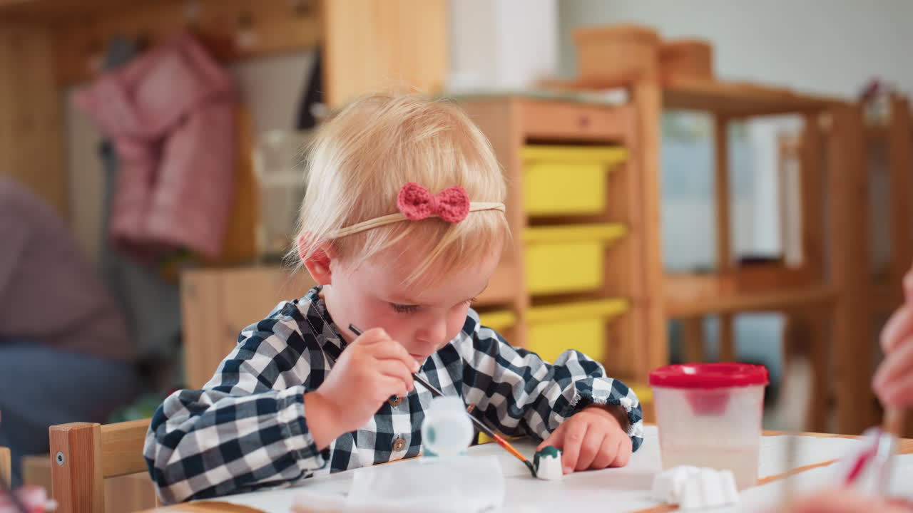 Kindergarten girl with blonde hair wearing checkered shirt and pink bow headband holds brush while painting small plaster figurine during classroom art session with other children