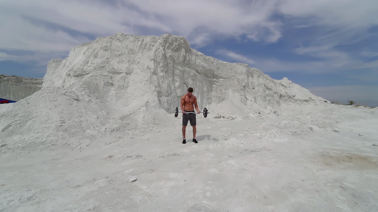 Sporty man lifting heavy barbell in the mountains. Bodybuilder doing his workout on the white background of a rocky hill. Slow motion.