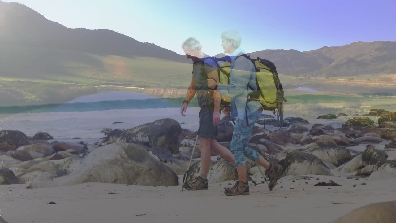 Senderistas maduros entrando en escena, caminando por la orilla rocosa al amanecer, con mochilas amarillas y bastones de trekking para el viaje.