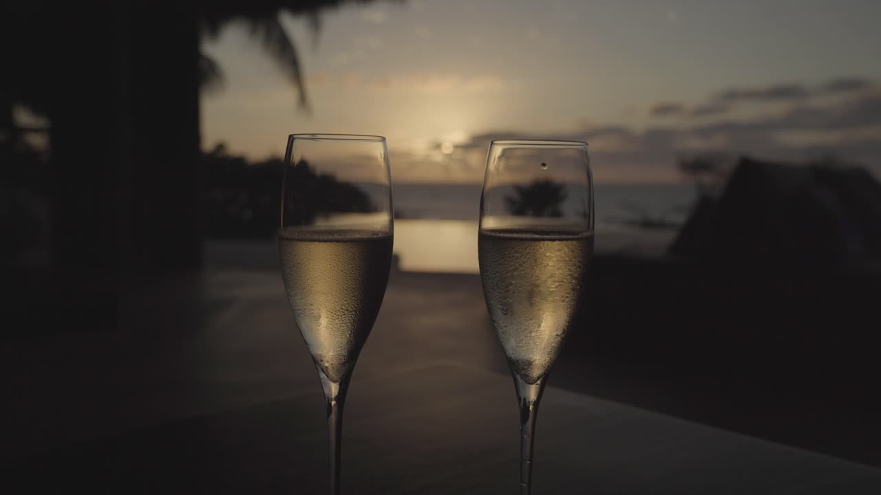 timelapse de copas de champán refrigeradas en una villa de lujo con una piscina infinita, océano y puesta de sol en el medio en el fondo