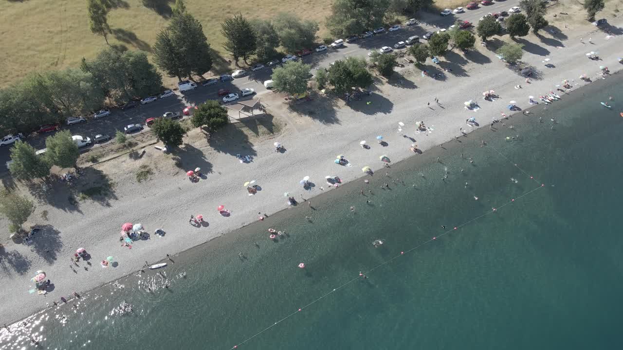 toma aérea en el lago ranco en chile de gente bañándose y divirtiéndose en el lago con autos estacionados cerca de un campo de hierba con árboles