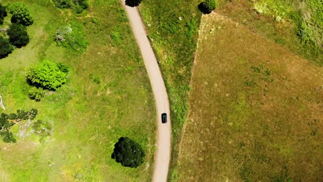 Vista-Aérea-De-Pájaro-Siguiendo-Un-Coche-Negro-Que-Viaja-A-Lo-Largo-De-Una-Pequeña-Carretera-Rural