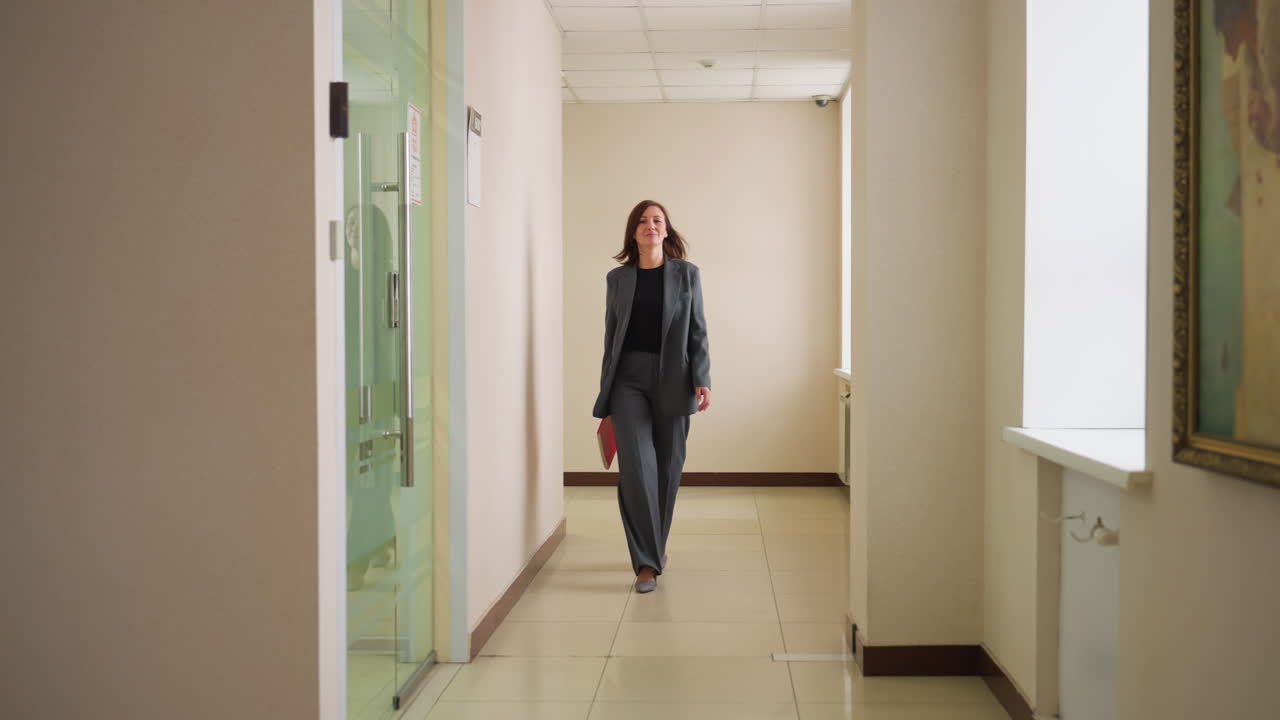Confident businesswoman walking down office hallway in formal suit holding red notebook, heading toward meeting room with glass door, bright modern corporate interior, professional atmosphere