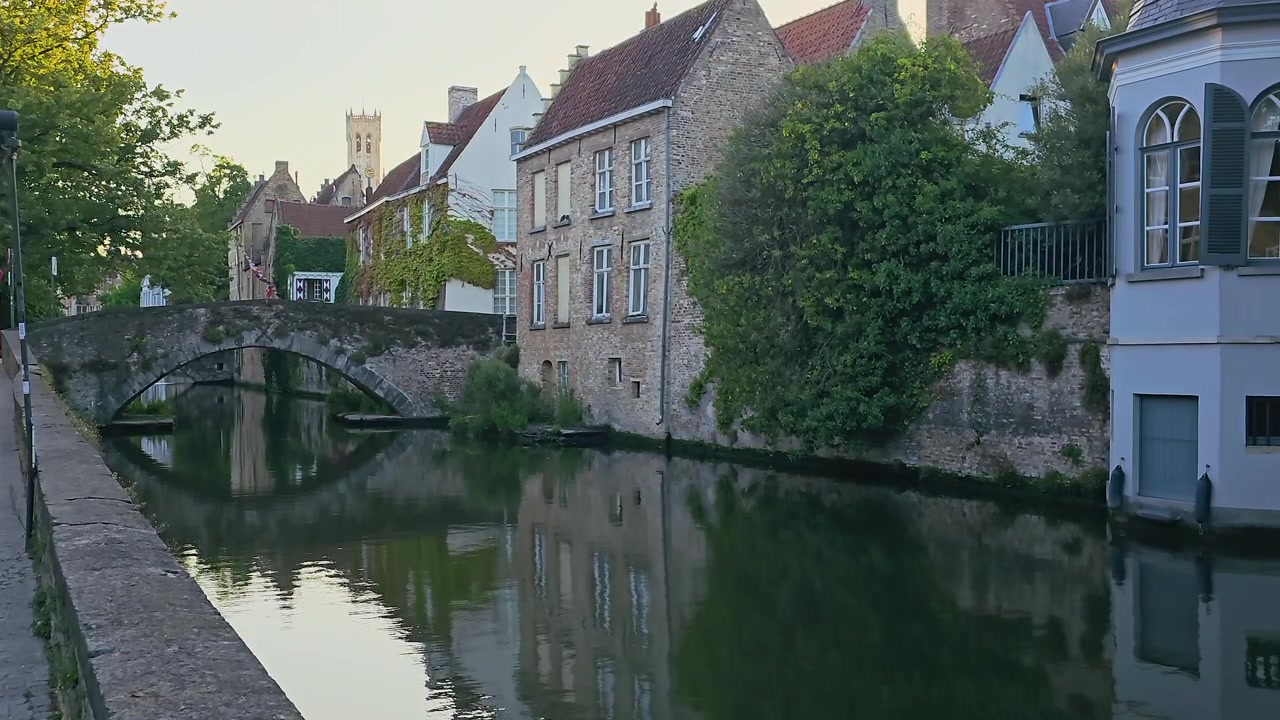 Heidelberg, Tyskland, havnefrontsarkitektur langs en rolig kanal med middelalderlige murstensbygninger, en stenbro, efeu-dækkede facader, hængende blomster og fredfyldte refleksioner af den gamle bydel ved solnedgang