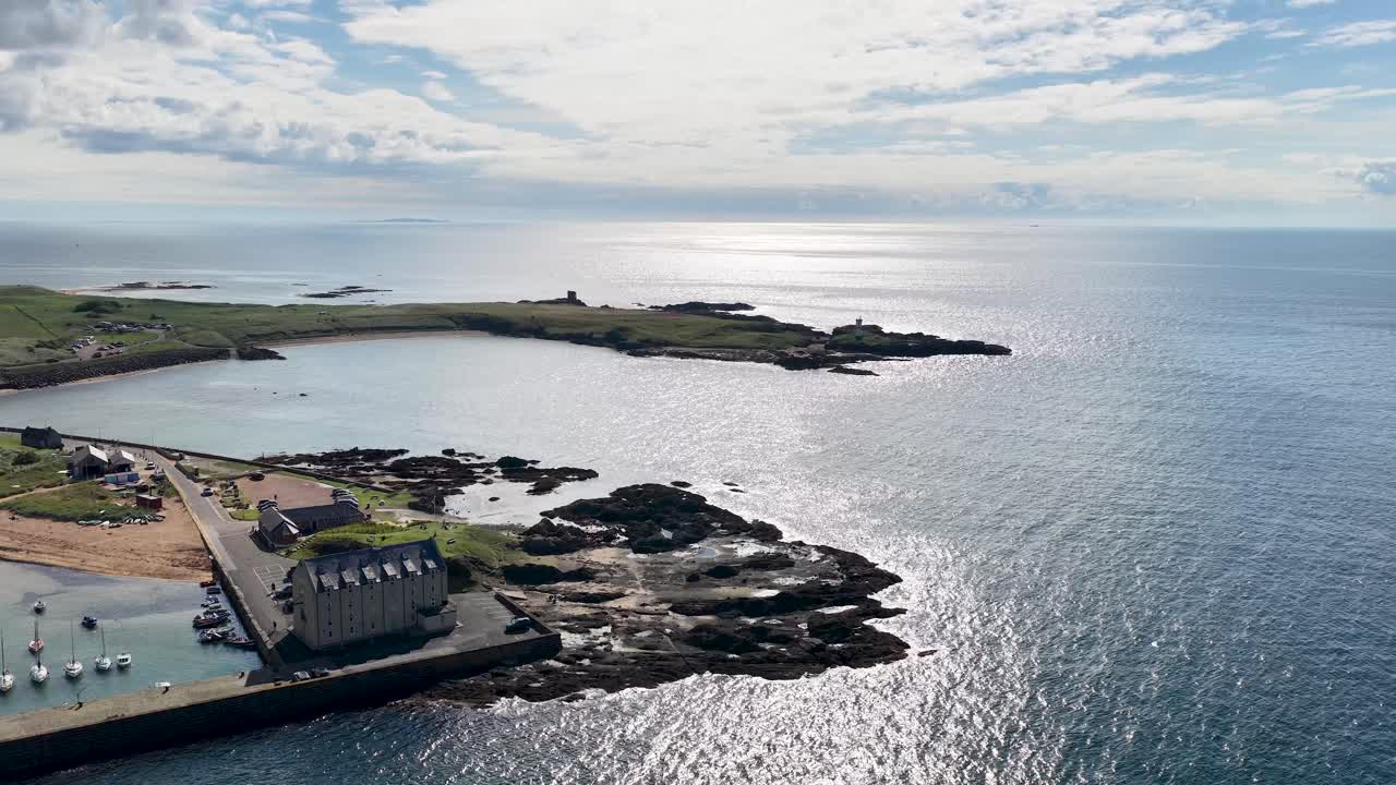 Drone glides above Fife harbor, lighthouse ruins, sparkling water, and seaside village in daylight