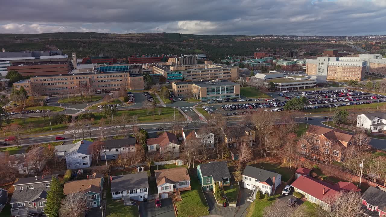 Aerial panorama of a hospital or campus complex with modern buildings and packed car parks, surrounded by roads and hills under a cloudy sky