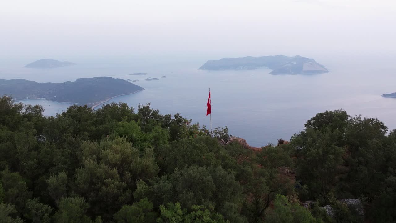 Kaş, Antalya sahil kasabasına bakan yemyeşil tepeler üzerinde havadan dolly çekimi, dağdaki bir direkte Türk bayrağıyla genel bir görünüm sunuyor.