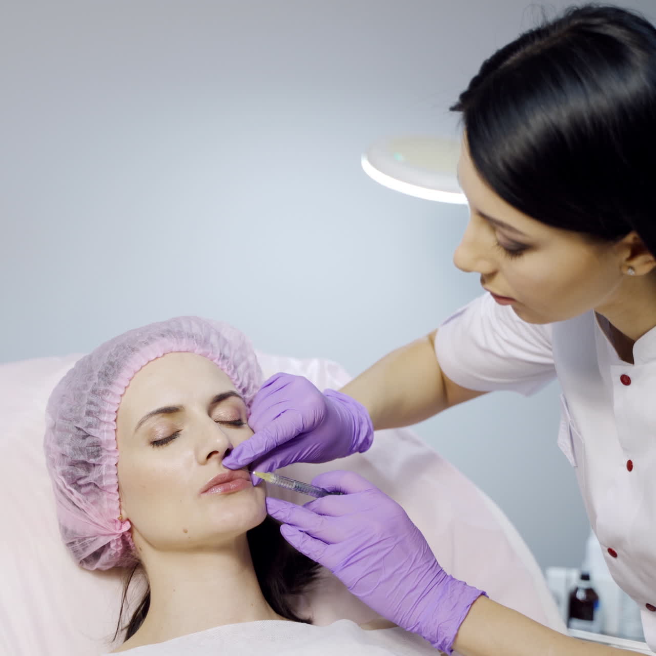 A cosmetologist in a white lab coat performs procedure of lip augmentation for a client in a beauty salon on a light background. Modern methods of cosmetology.