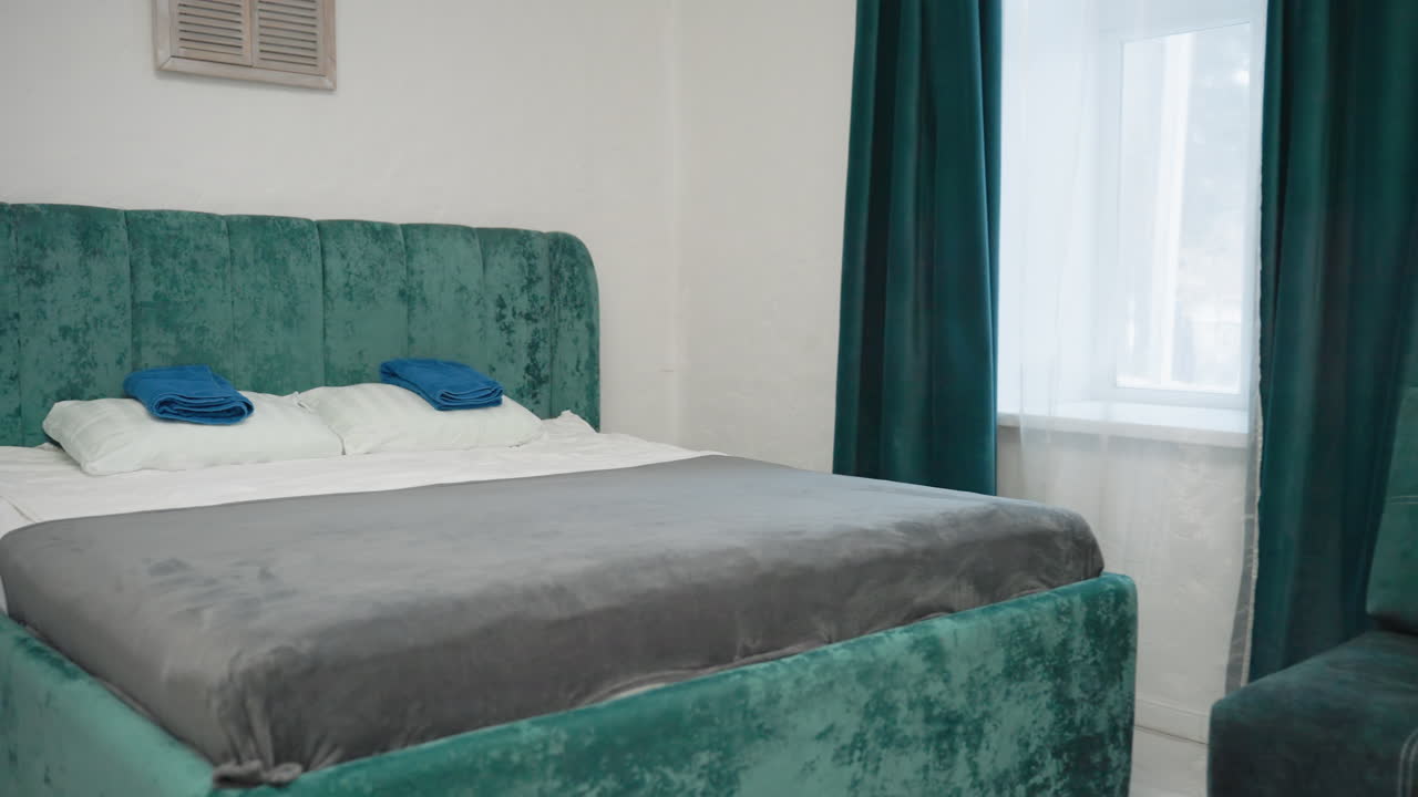 Modern bedroom with green velvet bed frame, plush gray bedding, teal curtains, and cozy green velvet couch with cushions. Bright natural light coming through large window