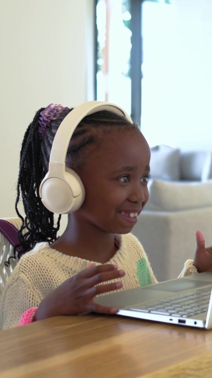 Vertical video: Waving girl in cream knit answering call at home using white headphones and laptop