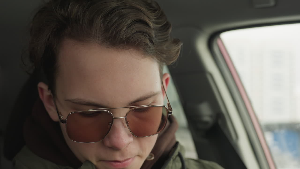 close up of young man in green winter jacket adjusting seatbelt inside car with focused expression as natural light filters through side window showing cold weather and blurred exterior cityscape