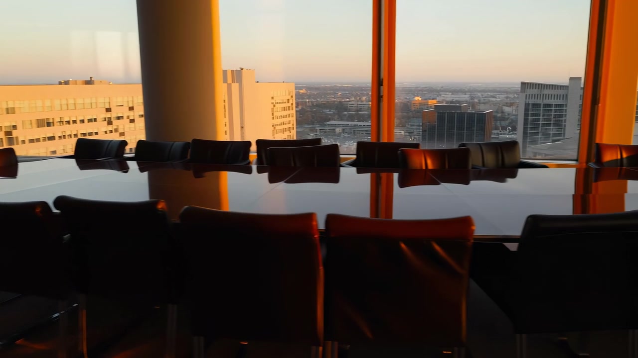 Der Innenraum eines leeren Konferenzraums zeigt dunkle Stühle, eine reflektierende Tischoberfläche und große Fenster mit entfernten Gebäuden draußen, während warmes Sonnenlicht eine ruhige Arbeitsplatzatmosphäre und eine großzügige Raumaufteilung schafft
