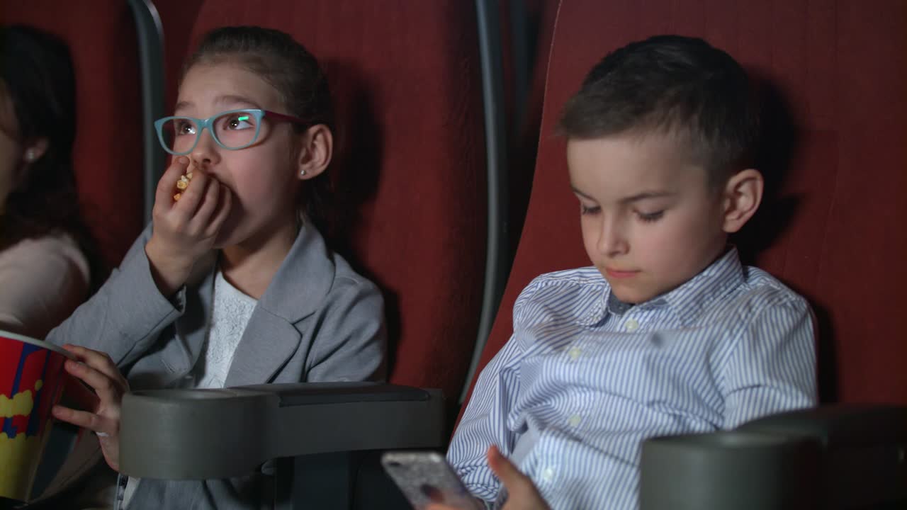 una niña comiendo palomitas de maíz en el cine. un niño usando un teléfono inteligente en el cine