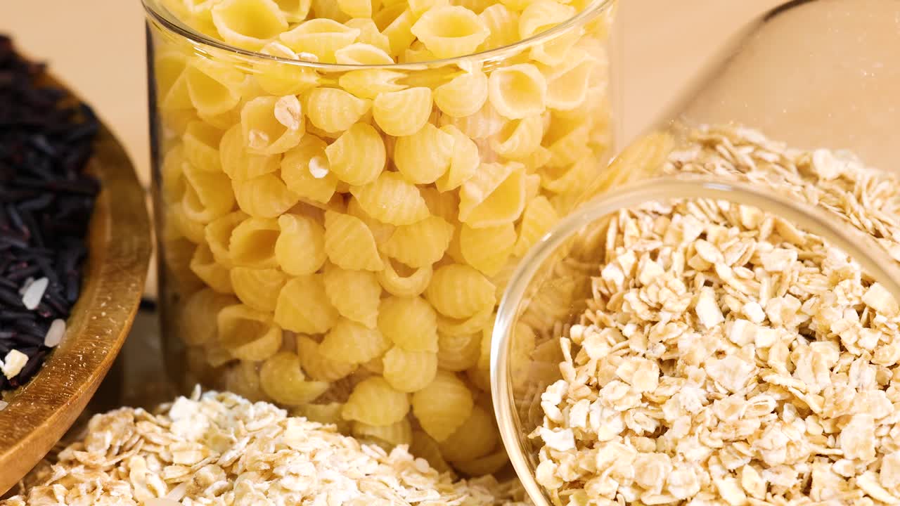 Close-up of rice, pasta, and oats in jars on a wooden surface. Bright lighting highlights textures and colors