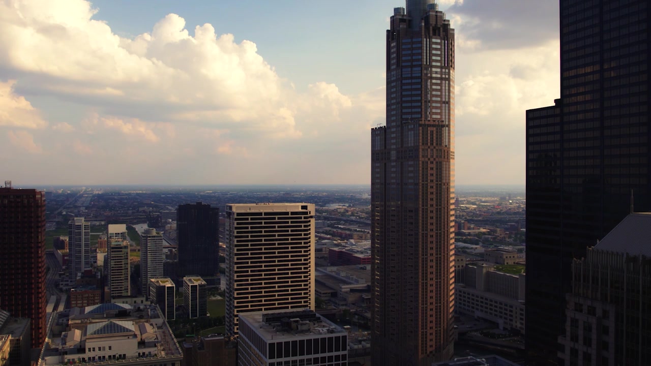 Veduta aerea con drone di un'alta torre nel centro di Chicago, circondata da una fitta architettura cittadina, nuvole drammatiche e la calda luce dell'ora d'oro.