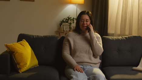 young woman spending evening at home sitting on sofa talking on mobile phone