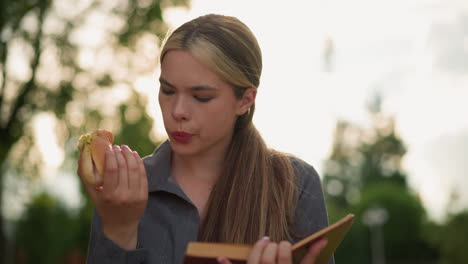 lady in grey top examines snack in hand while eating and reading book outdoors, seated on park bench surrounded by trees and greenery with blurred background, soft sunlight illuminates