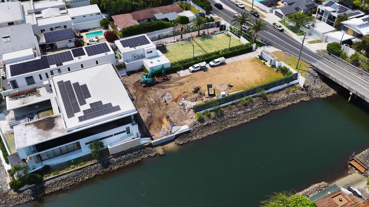Drone aerial pans over waterfront construction site beside modern homes, canal, bridge, and parked vehicles