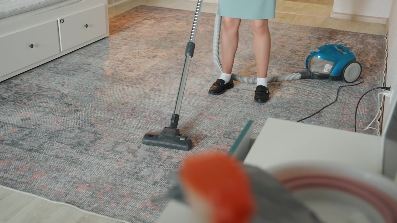 Detailní záběr nohou vysávajících koberec v domě. Bílá žena ve světle modrých šatech provádí důkladné čištění koberce s modrým sáčkovým vysavačem, zaměřeno na hlavici vysavače a nohy, texturovaný koberec, úložné zásuvky, jemné světlo.