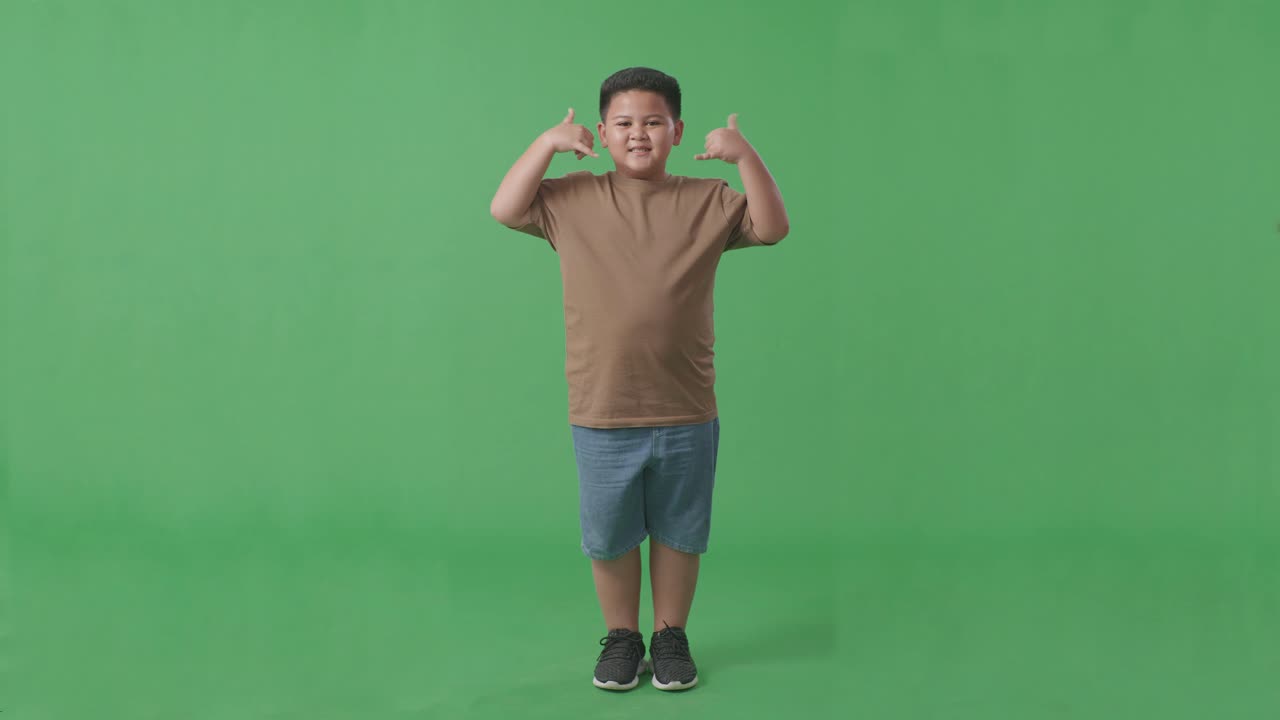 카메라에 미소 짓고 녹색 스크린 배경 스튜디오에 서있는 동안 나를 호출 제스처를 만드는 아시아 소년의 몸으로 가득 차 있습니다.
