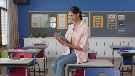 in school, woman sitting on desk using tablet in classroom, smiling