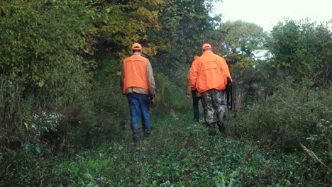 three men in camouflage and blaze orange carrying rifles while hunting in the bush