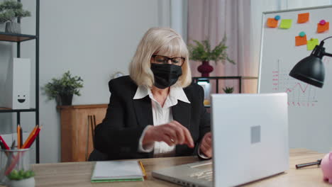 Senior-employee-wears-face-mask-works-alone-from-home-office-sits-at-desk-using-laptop-computer