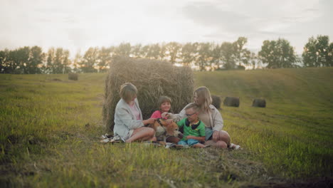 joyful family seated on grass near hay bale, enjoying quality time outdoors while petting their dog, warm sunlight casts a golden glow, creating a heartwarming and peaceful rural atmosphere