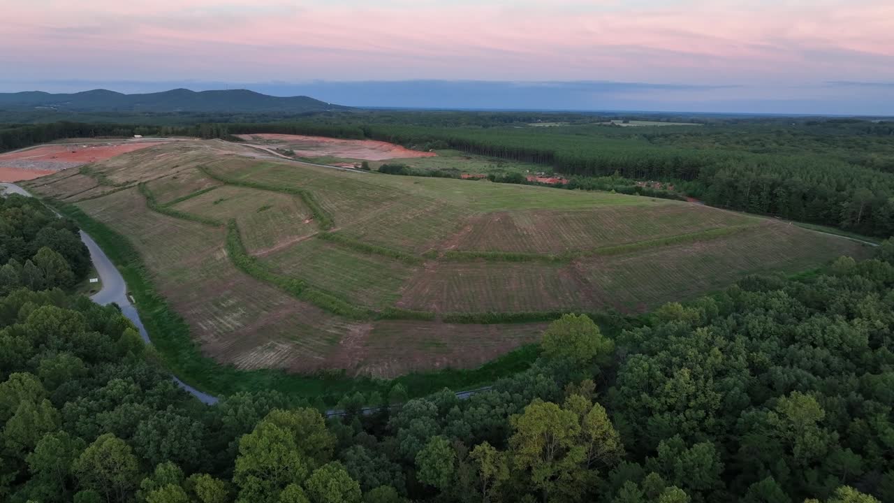 Aerial view of serene landscape featuring rolling hills and expansive fields. Winding road bordered by lush greenery, leading into a tranquil environment. Sunset time in America. Wide shot