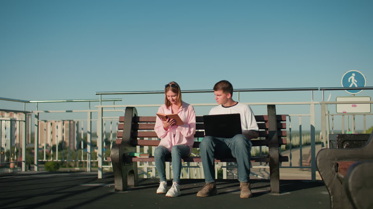 남자와 여자 학생들이 벤치에 앉아 함께 공부하고, 소년은 노트북에 타이핑하고 소녀가 책에서 읽는 것을 듣고, 동의 표시로 고개를 숙이고, 도시 배경에 배치됩니다.