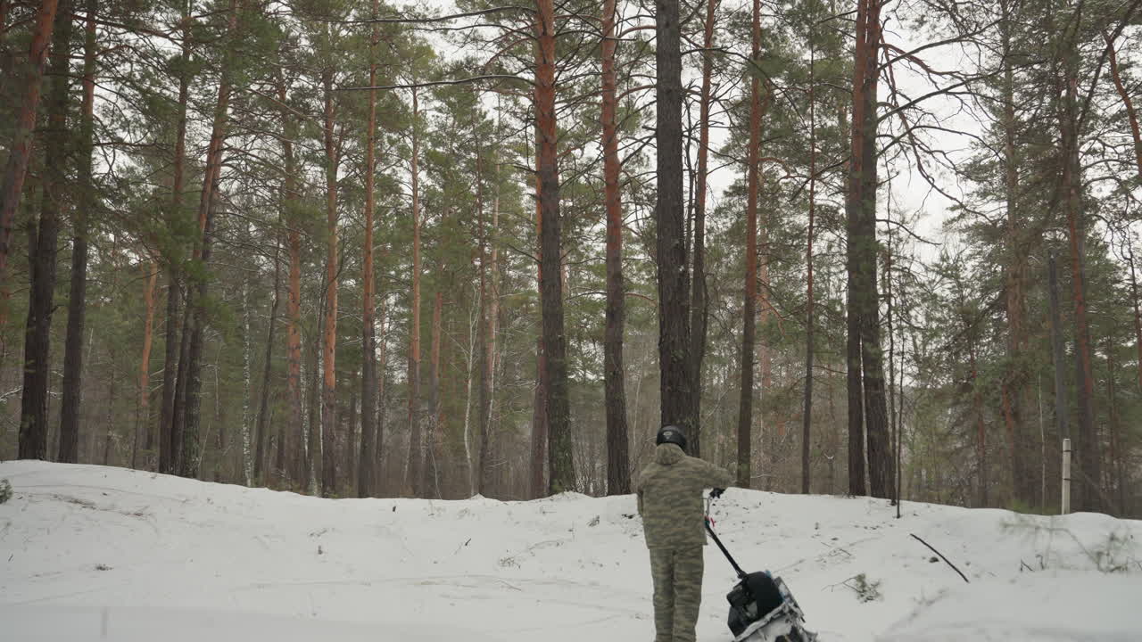 Guidatore con quad nella foresta innevata che ispeziona il quad cingolato in panne vicino a pini alti, leggera nevicata e luce invernale soffusa creano una scena di recupero tranquilla e tesa con tracce visibili e equipaggiamento imballato.