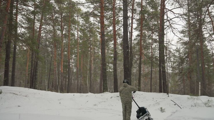 Guidatore con quad nella foresta innevata che ispeziona il quad cingolato in panne vicino a pini alti, leggera nevicata e luce invernale soffusa creano una scena di recupero tranquilla e tesa con tracce visibili e equipaggiamento imballato.