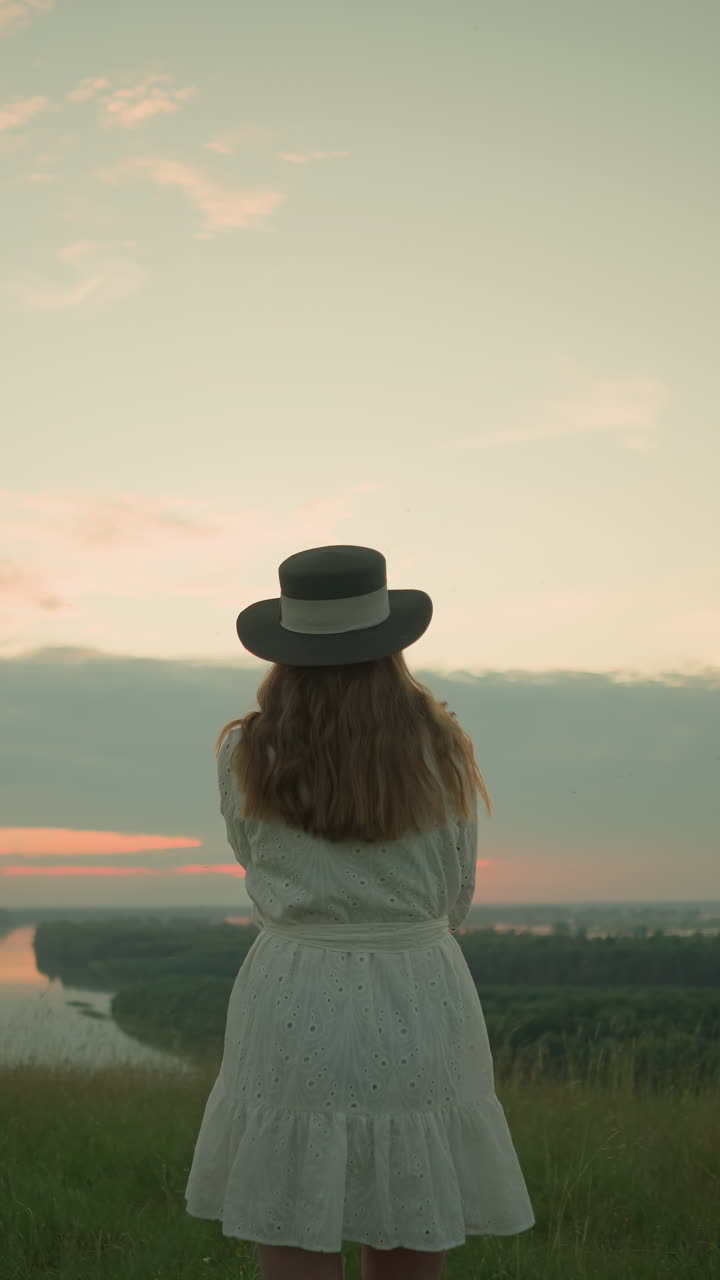 Back view of an adult woman in a white dress and hat standing alone in a serene grassy field, gazing out over a tranquil lake during sunset under a soft, pastel-colored sky