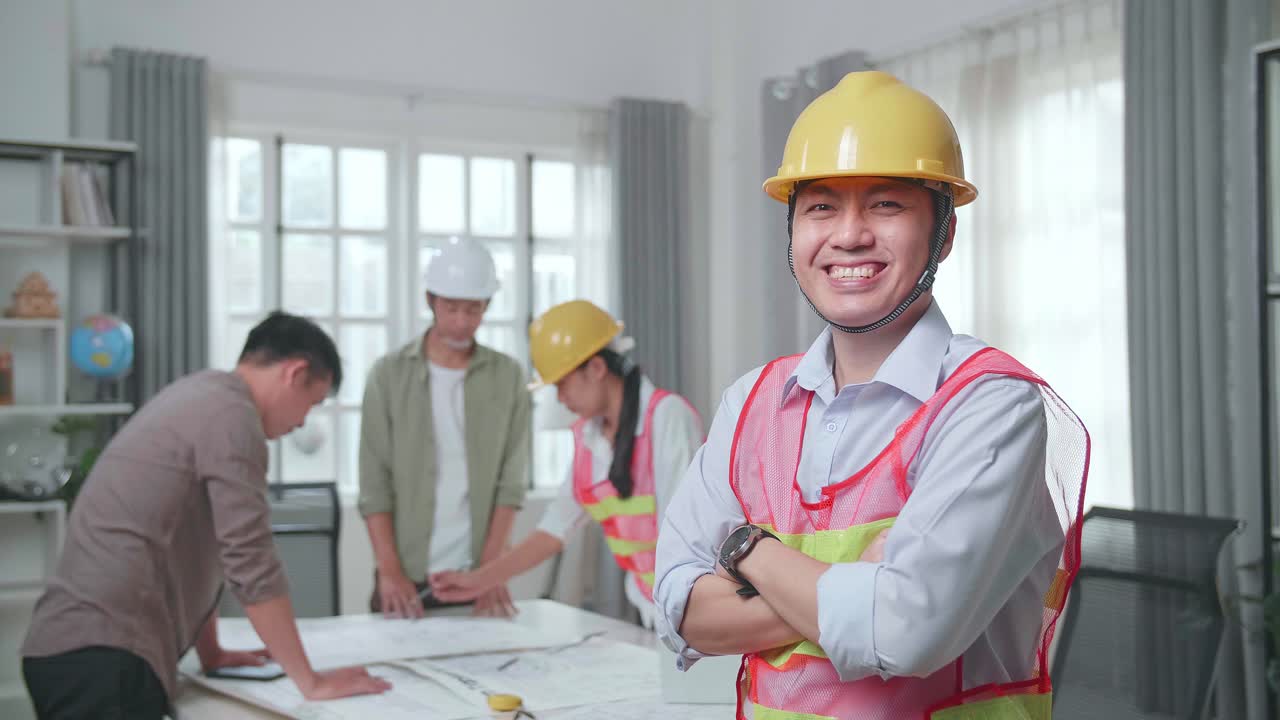 Asian Man Engineer With Helmet Crossing Arms And Smiling To Camera While His Colleagues Are Discussing About Work With A Man At The Office