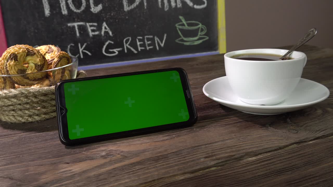 bebida caliente, galletas y teléfono inteligente en la mesa en el café. teléfono inteligente pantalla verde.