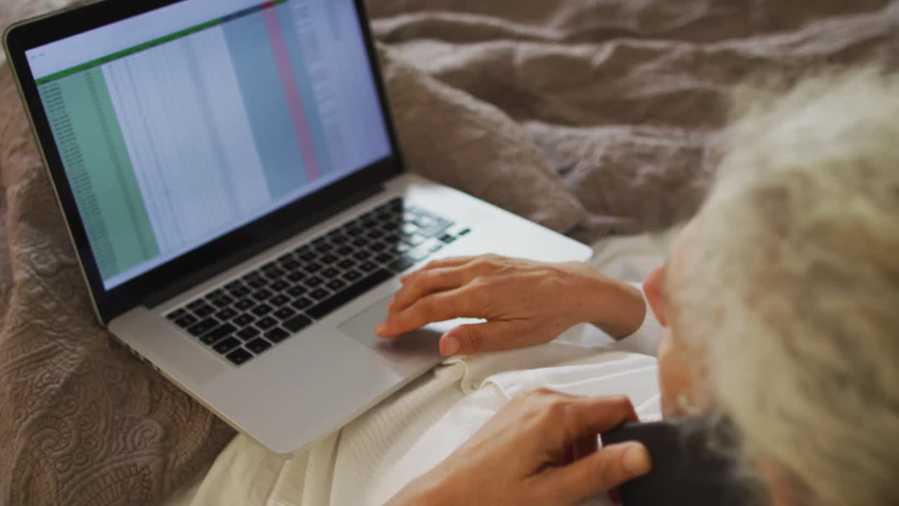 Senior mixed race woman lying in bed using laptop and talking on smartphone