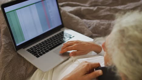 senior mixed race woman lying in bed using laptop and talking on smartphone