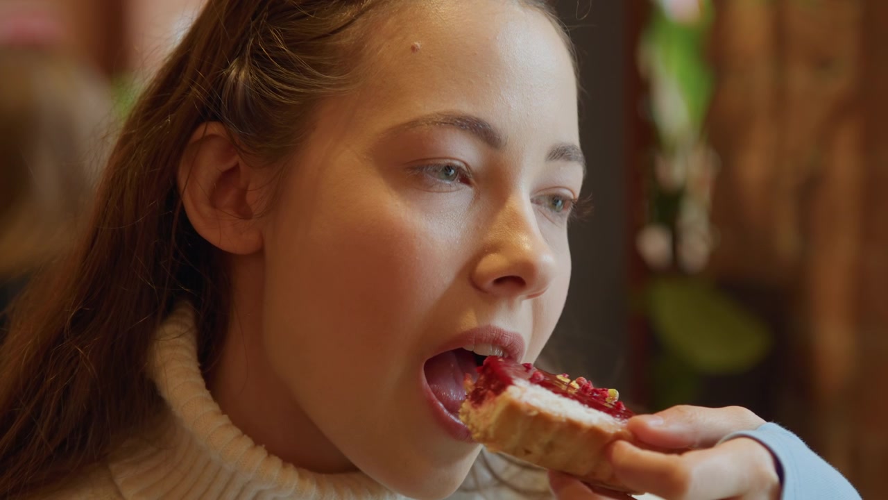 Nærbilde av en ung kvinne som spiser kake i mykt lys. Kvinnen holder og tar biter av desserten, med varme innendørs bakgrunnsdetaljer synlige overalt. Scenen fokuserer på hennes handlinger.