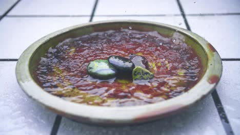 Cierre-De-Cámara-Lenta-De-Lluvia-Que-Inunda-Una-Jarra-De-Arcilla-De-Flores-Marrones-Con-Azulejos-Blancos-En-El-Fondo-Y-Piedras-Escritas-Con-Las-Palabras-Calma-Y-Suerte