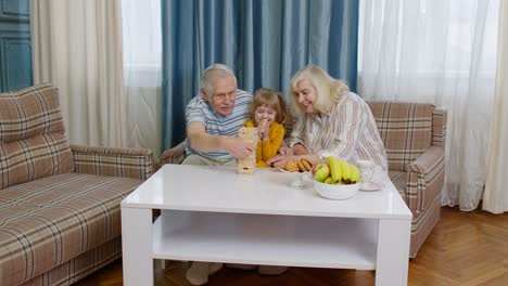 Pareja-De-Ancianos,-Abuelos-Y-Nieta-Disfrutando-De-Un-Juego-De-Mesa-Construyendo-Una-Torre-A-Partir-De-Bloques-En-Casa