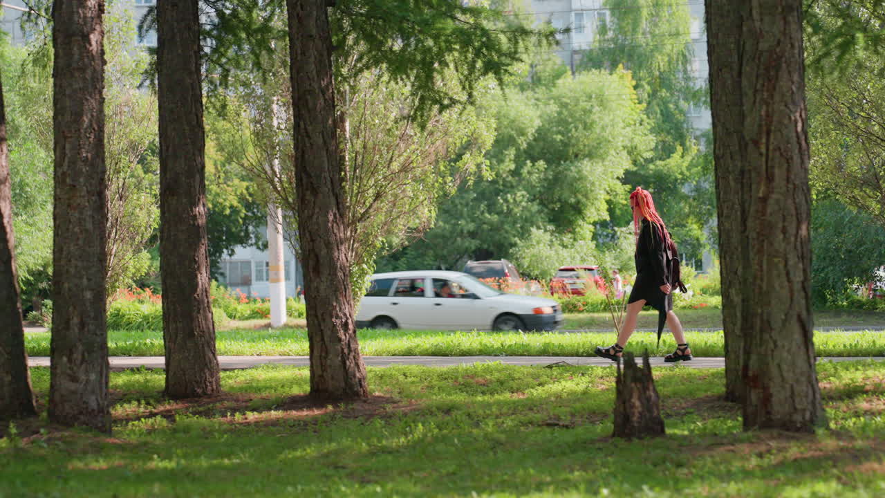 Спокойная женщина проходит мимо придорожной рощи, женщина европеоидной расы прогуливается среди высоких деревьев, безмятежная сцена; женщина европеоидной расы идет между высокими стволами возле припаркованных автомобилей в тихой обстановке.