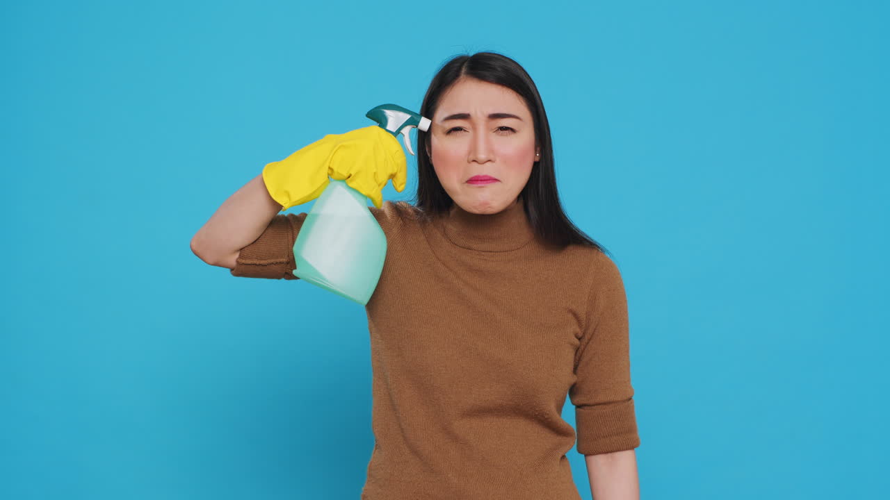 Upset angry maid putting detergent spray in front of temple while pretending to shoot herself after finishing cleaning