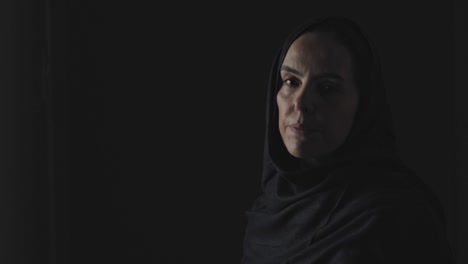 a muslim woman in a somber mood turns from darkness to gaze at the camera